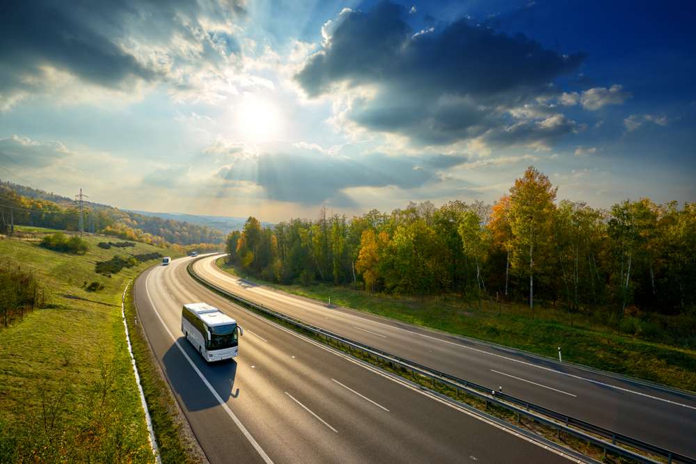 Three white buses traveling on the asphalt highway between deciduous forest in autumn colors under the radiant sun and dramatic clouds. View from above.