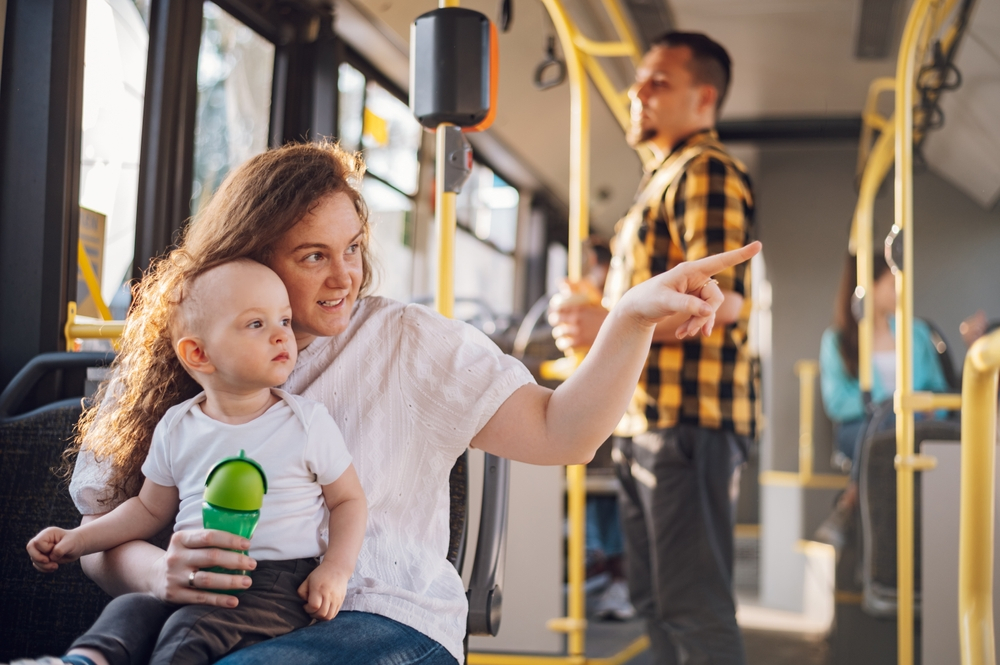 Young plus size mother in casual wear holding little toddler boy in her lap and together looking at window. Mom and her child enjoying a bus journey together. Mother pointing her finger. Copy space.